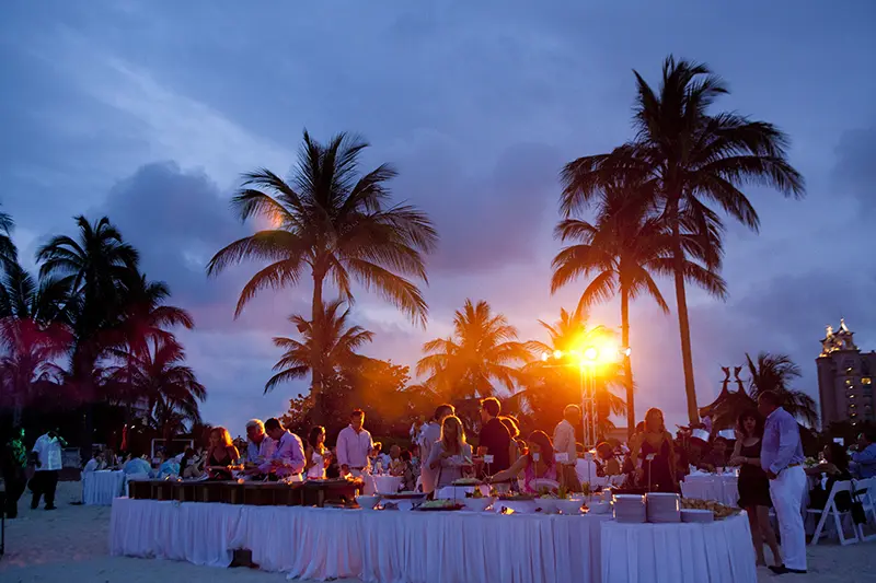 Outdoor private celebration at sunset with palm trees, dining tables, and a waterfront setting.